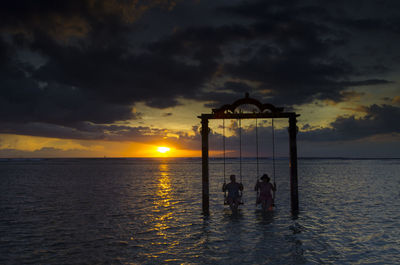 Silhouette couple swinging in sea against cloudy sky during sunset