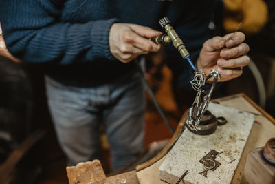 Anonymous goldsmith using blowtorch to heat tiny metal ornament while making jewellery on workbench