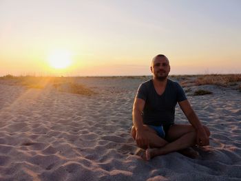Portrait of mid adult man sitting on beach against sky during sunset