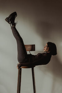 Side view of slim female in black outfit balancing on wooden stool with raised legs in room against white wall