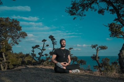 Young man sitting on tree against sky