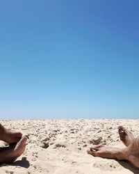 Low section of man lying on sand at beach against clear blue sky