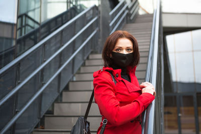 A young woman wearing a black protective medical mask near a glass building with a staircase 