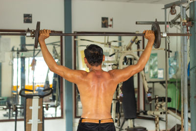 Rear view of shirtless man exercising with dumbbell at gym