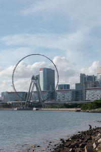 View of ferris wheel by buildings against cloudy sky