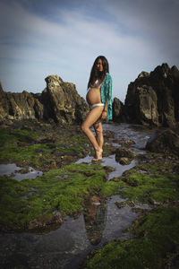 Pregnant young woman standing on rock at beach against sky