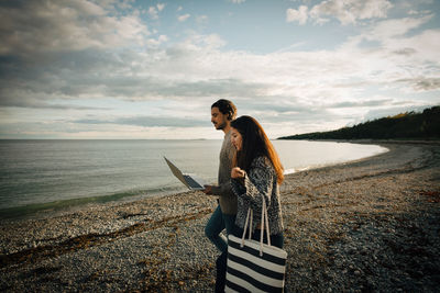 Woman walking with man using laptop on sea shore at beach against sky