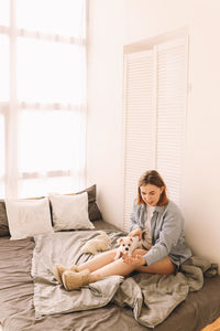 A cheerful young teenage woman plays with her pet a small dog in bed in the morning in a cozy house