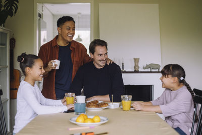 Happy gay couple having fun while having breakfast with daughters at dining table in home