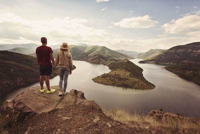 Rear view of couple standing against river and mountains on cliff