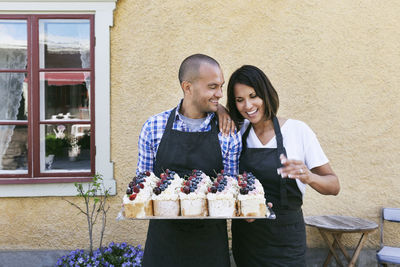 Smiling bakers holding tray of pastries while standing against wall outside cafe