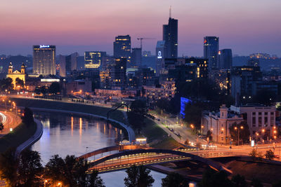 Illuminated bridge over river by buildings against sky at night