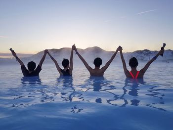 Friends with arms raised in hot spring against sky at sunset