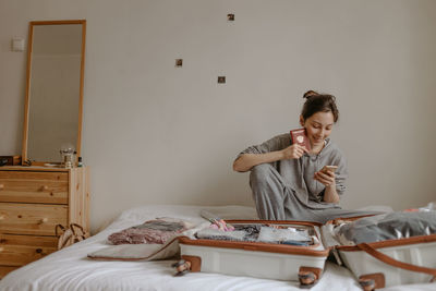 Woman checking list before going to airport, passport in her hand and open suitcase on the bed