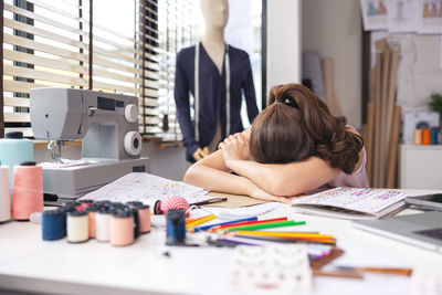 Young exhausted seamstress with a measuring tape around her neck, sitting at an electric sewing 