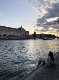 Women sitting on riverbank against sky during sunset