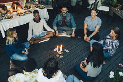 Multi-ethnic male and female friends sitting with eyes closed around lit candles and mobile phones in balcony during gro