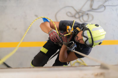 Overhead view of young fireman in protective hardhat and gloves ascending wall on colorful rope during routine practices
