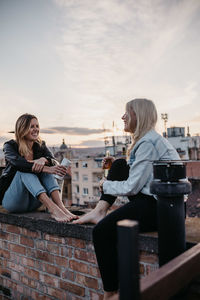 Cheerful women talking while sitting on building terrace against sky