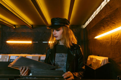 Tranquil young female in black cap choosing vinyl record while standing near counter with collection of musical albums in store with glowing lamps