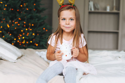 Cute smiling funny little girl in pink dress drinking cocoa on bed in room with christmas tree