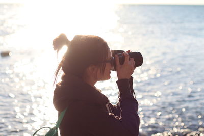 Side view of woman photographing with camera by sea during sunny day