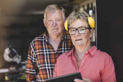 Portrait of confident senior woman wearing ear protectors while standing with man at workshop doorway