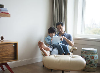 Father and son using tablet while sitting against window at home