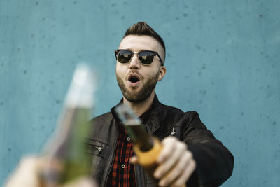 Portrait of young man wearing sunglasses holding beer bottle against wall outdoors