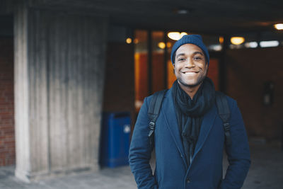 Portrait of happy man wearing blue warm clothing and knit hat standing in campus