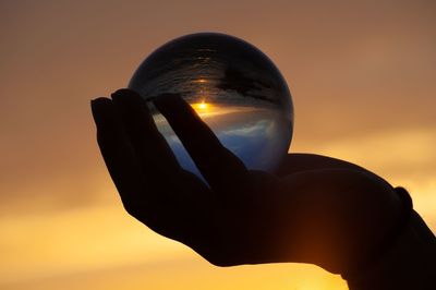 Silhouette hand holding crystal ball against sky during sunset