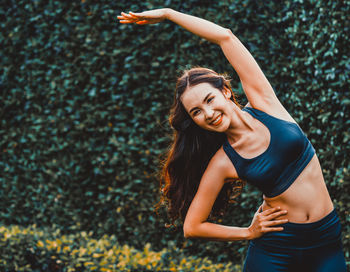 Smiling young woman exercising while standing against plants