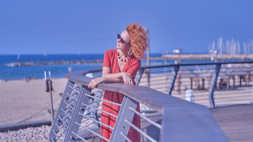 Woman sitting on railing at beach against sky