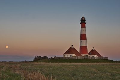 Lighthouse on field by building against sky during sunset