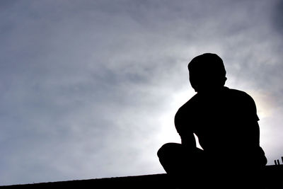 Low angle view of silhouette boy sitting against sky