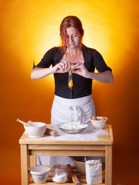 Woman preparing food with egg while standing against orange background