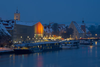 Illuminated buildings by river against sky at sunset