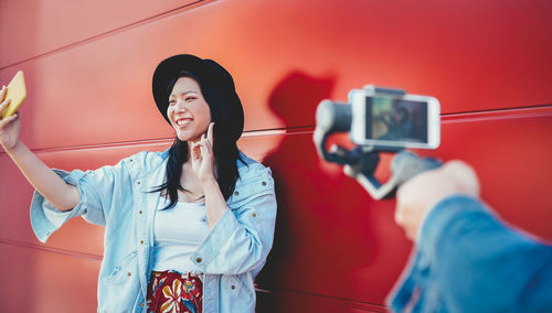 Smiling young woman gesturing peace sign while taking selfie against red wall