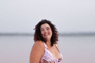 Delighted curvy female in bikini standing near water of pink pond in summer and looking at camera