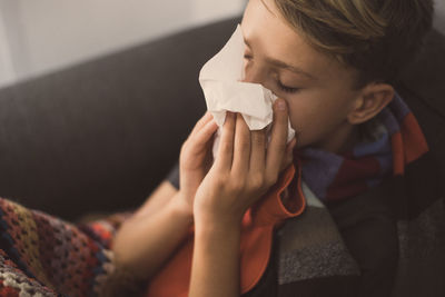 Close-up of boy blowing nose while sitting on sofa at home