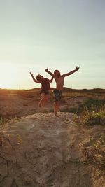 Two children jumping on beach against sky during sunset