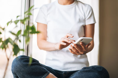 Young brunette woman in casual clothes using mobile phone sitting on window sill at home