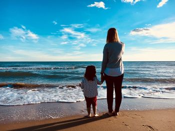 Rear view of woman with daughter standing on shore at beach against sky