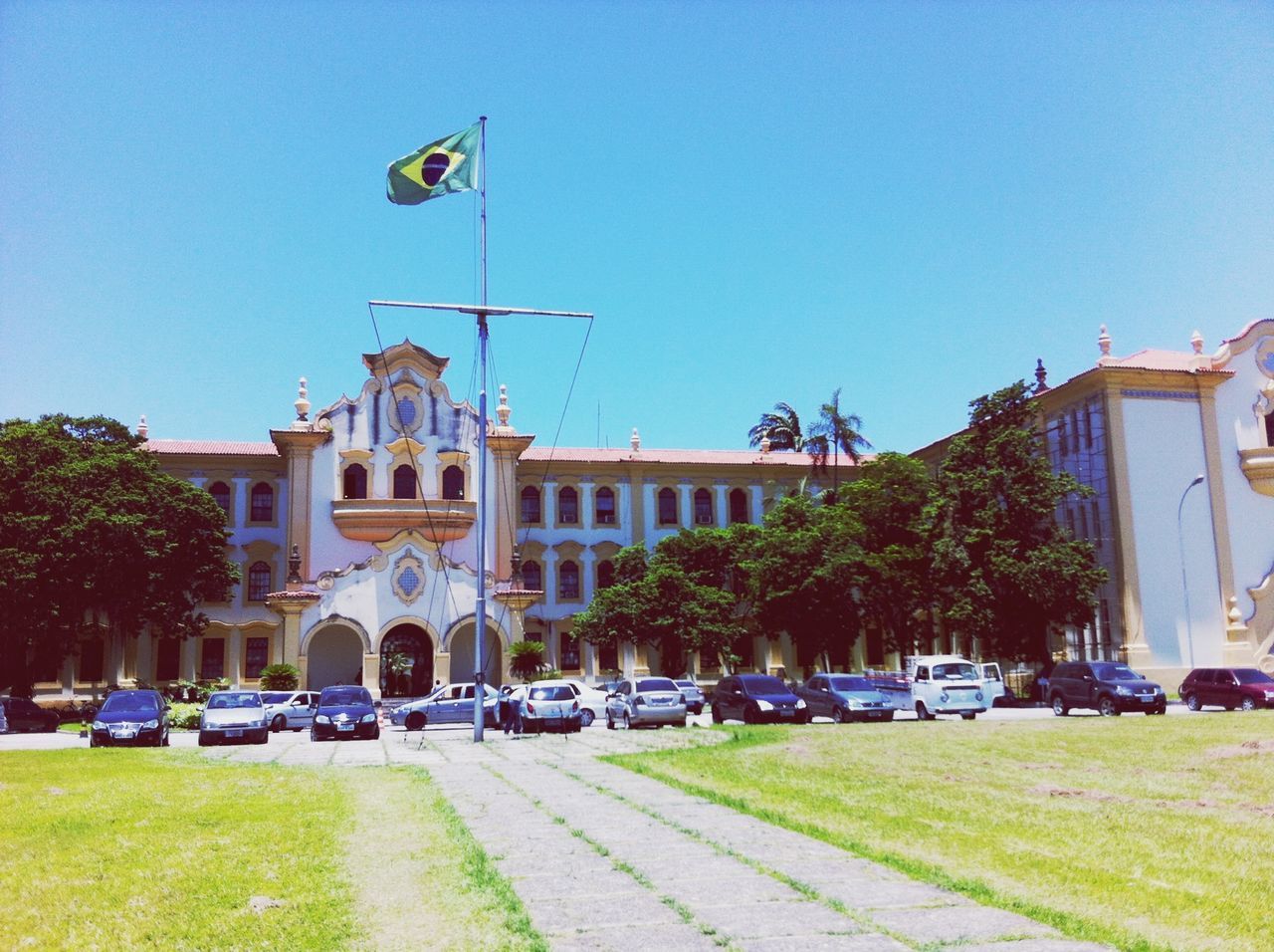 building exterior, car, transportation, land vehicle, architecture, mode of transport, built structure, clear sky, street, tree, road, city, grass, blue, parking, sunlight, city street, stationary, incidental people, day