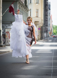 Portrait of young woman holding wedding dress and walking on street