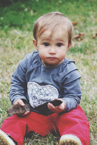 Portrait of cute baby  with widely opened eyes sitting on grass field holding wooden sticks in hands