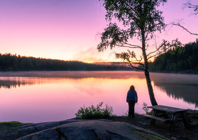 Rear view of woman standing at lake against sky during sunset