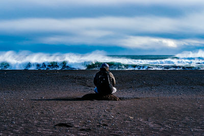 Rear view of backpacker sitting at beach against cloudy sky