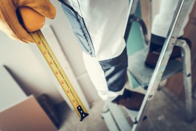 Low section of worker holding tape measure while standing on ladder at home