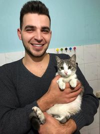 Portrait of smiling young man carrying cat while standing against wall at home